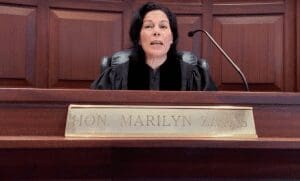 Court of Appeals Judge Marilyn Zayas as she takes a seat at the bench of a courtroom, behind a plaque reading 'Hon. Marilyn Zayas.'